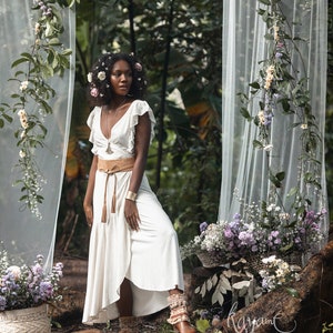 Puede incluir: Una mujer con un vestido blanco con un cintur&oacute;n marr&oacute;n y una corona de flores se encuentra en un entorno de bosque. Est&aacute; rodeada de vegetaci&oacute;n y flores. El vestido tiene una falda fluida y un escote con volantes. La mujer mira hacia un lado.