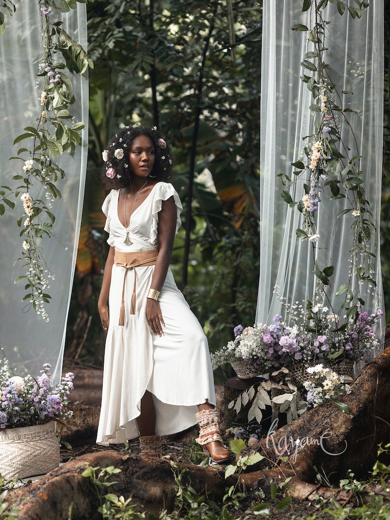 Puede incluir: Una mujer con un vestido blanco con un cintur&oacute;n marr&oacute;n y una corona de flores se encuentra en un entorno de bosque. Est&aacute; rodeada de vegetaci&oacute;n y flores. El vestido tiene una falda fluida y un escote con volantes. La mujer mira hacia un lado.