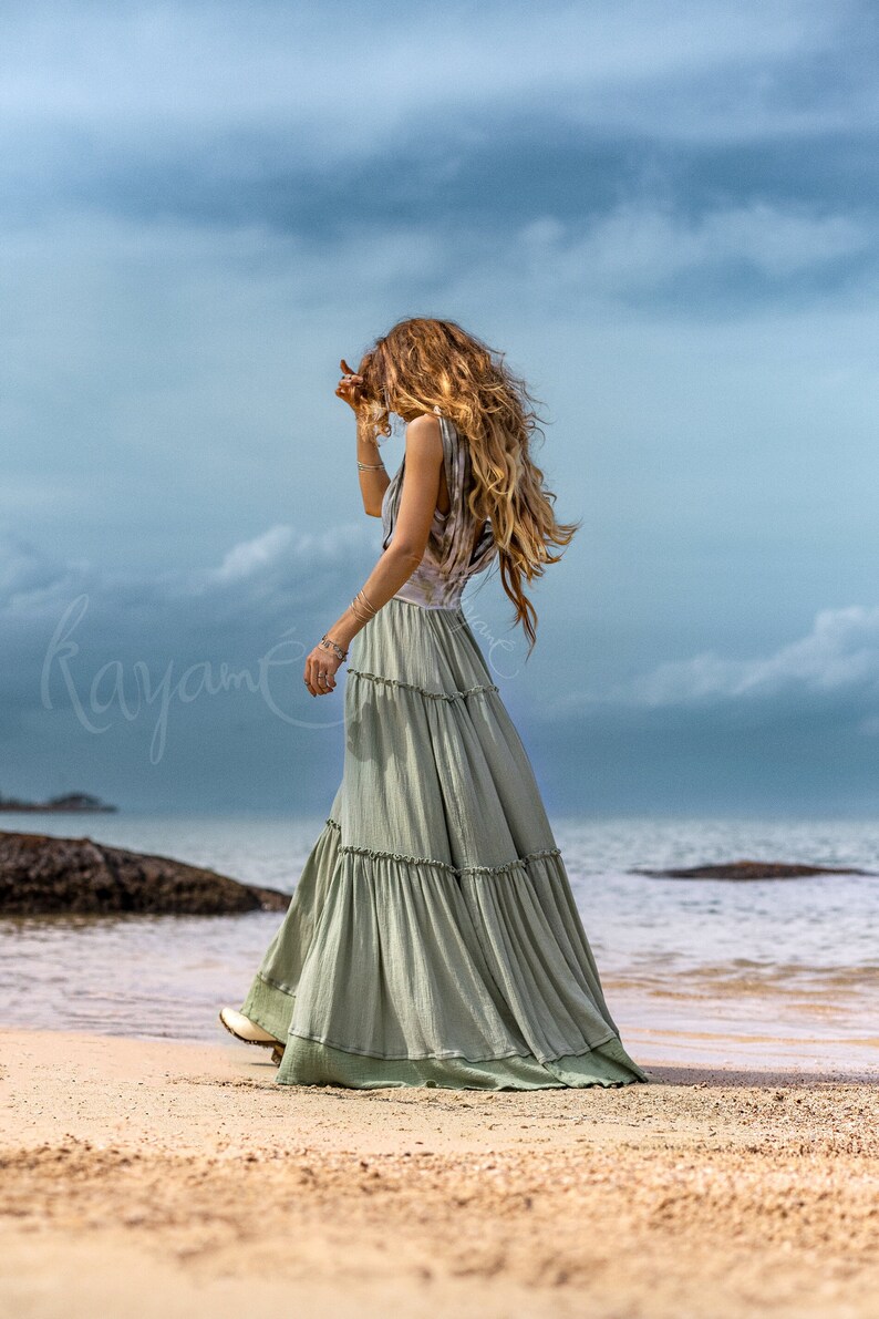 Puede incluir: Una mujer con un vestido maxi verde claro con m&uacute;ltiples niveles y volantes, de pie en una playa de arena con el oc&eacute;ano de fondo.
