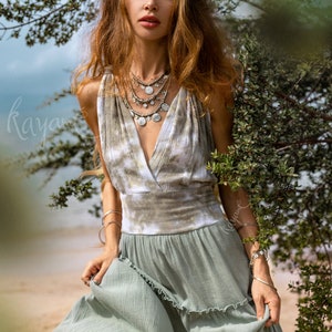 Puede incluir: Una mujer con un vestido maxi verde con un corpi&ntilde;o de te&ntilde;ido anudado y collares de plata superpuestos. Est&aacute; de pie frente a un arbusto verde frondoso.