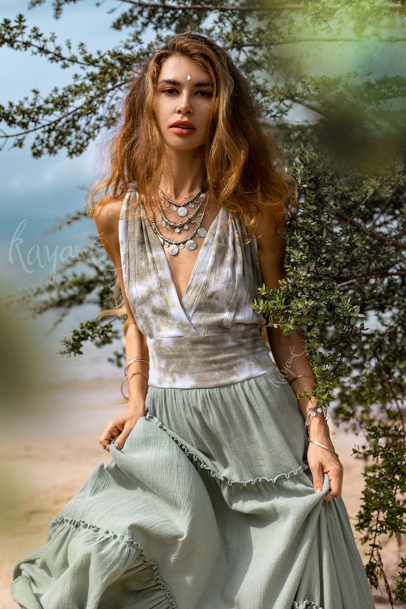 Puede incluir: Una mujer con un vestido maxi verde con un corpi&ntilde;o de te&ntilde;ido anudado y collares de plata superpuestos. Est&aacute; de pie frente a un arbusto verde frondoso.