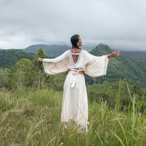 May include: A woman wearing a white, flowing, long-sleeved dress with a cinched waist and a deep V-neck. She is standing in a field of tall grass with a mountain range in the background.