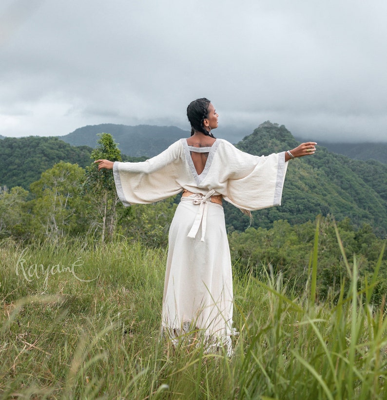 May include: A woman wearing a white, flowing, long-sleeved dress with a cinched waist and a deep V-neck. She is standing in a field of tall grass with a mountain range in the background.