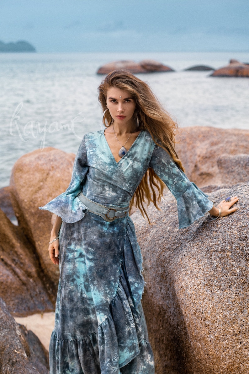 May include: A woman wearing a blue and white tie-dye wrap dress with a ruffled hem. She is standing on a rocky beach with the sea in the background.