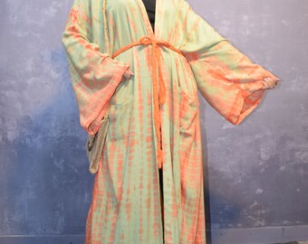 May include: A floor-length, open-front robe featuring a light green and orange tie-dye pattern, wide sleeves, and a matching orange fabric waist tie. The garment includes two front pockets and is worn with a dark wide-brimmed hat and a small bag.