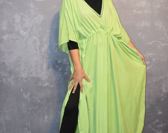 May include: A lime green kaftan dress featuring a V-neckline with dark decorative trim, short wide sleeves, and a drawstring waist. The garment is layered over a black long-sleeved top and black trousers, with side slits extending from the waist down.