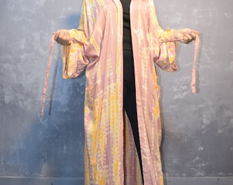 May include: A long, open-front kimono robe featuring a tie-dye pattern in shades of dusty pink and bright yellow. The garment has wide, loose sleeves, two front pockets, and matching fabric ties at the waist. A brown cowboy hat is worn above.
