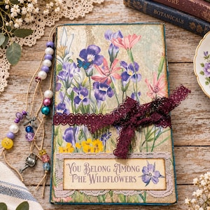 May include: A rectangular journal featuring a floral print with purple, pink, and yellow wildflowers. A dark maroon lace ribbon is tied around the center. A label at the bottom reads, "You Belong Among The Wildflowers." A beaded twine tassel hangs from the side.