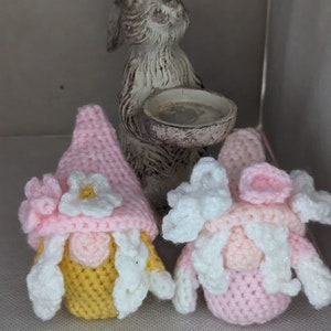 May include: Two crocheted gnomes with pink and white hats and yellow and white beards. The gnomes are sitting in front of a white ceramic rabbit figurine.