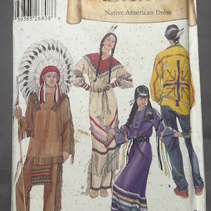 May include: A Simplicity Historic sewing pattern envelope 5446 for a Native American dress. The front features illustrations of four individuals wearing various fringed garments, including a tan outfit with a headdress, a cream dress, a purple dress, and a yellow shirt with dark pants.