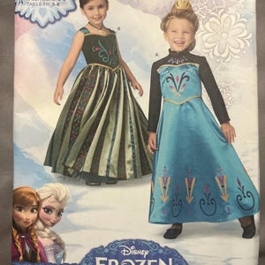 Puede incluir: Patrón de costura Simplicity 1222 para disfraces infantiles de Disney Frozen. El sobre muestra dos diseños de vestidos: uno verde oscuro con falda estampada y otro azul claro con mangas largas negras, ambos con coronas doradas y detalles florales.