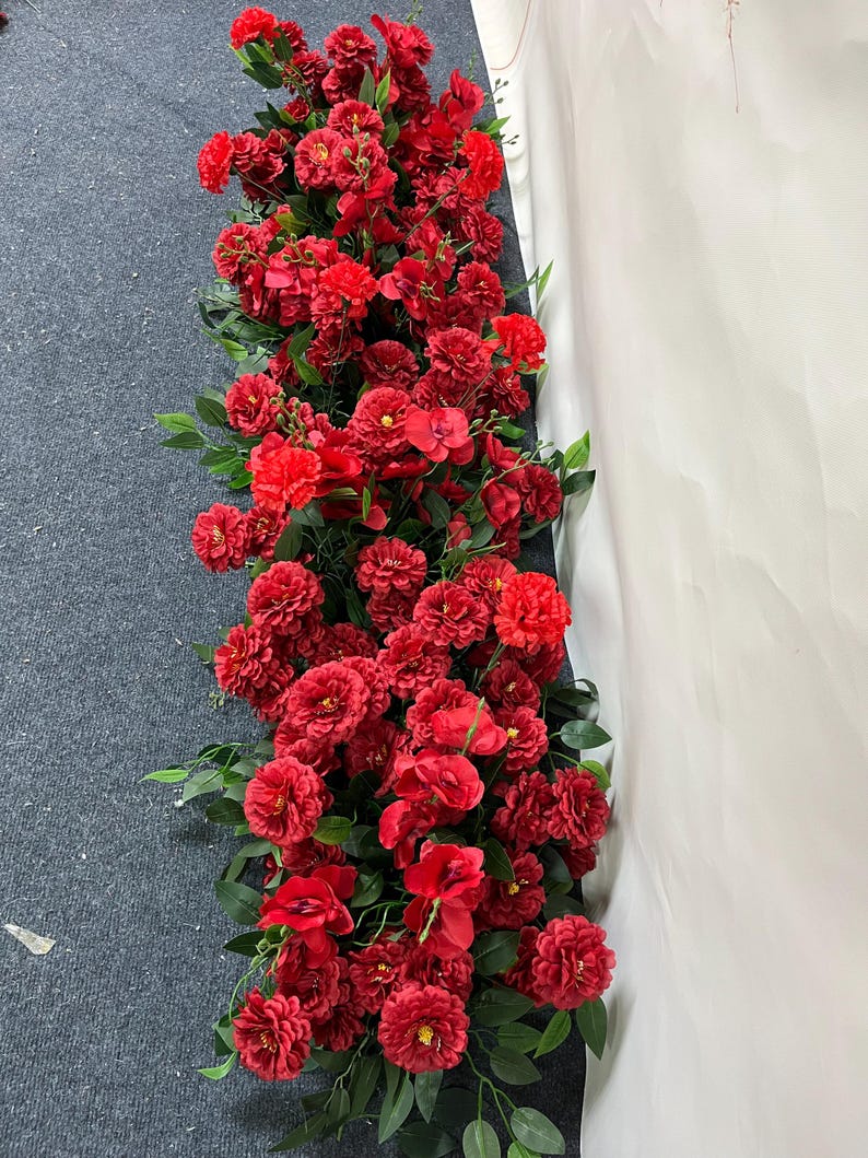 Puede incluir: Un arreglo denso de flores artificiales rojas, incluyendo rosas y otras flores, con hojas verdes. La exhibici&oacute;n floral es probablemente una guirnalda o guirnalda, adecuada para bodas o decoraci&oacute;n del hogar.