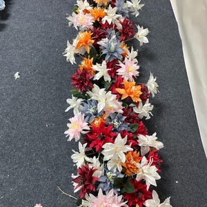 Puede incluir: Un vibrante arreglo floral con una mezcla de flores artificiales coloridas. El arreglo incluye flores blancas, naranjas, rojas, rosas y azules. Las flores est&aacute;n dispuestas en un patr&oacute;n lineal, adecuado para la decoraci&oacute;n de mesas o eventos.
