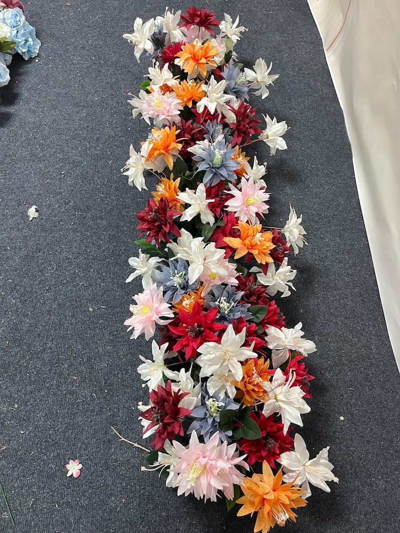 Puede incluir: Un vibrante arreglo floral con una mezcla de flores artificiales coloridas. El arreglo incluye flores blancas, naranjas, rojas, rosas y azules. Las flores est&aacute;n dispuestas en un patr&oacute;n lineal, adecuado para la decoraci&oacute;n de mesas o eventos.
