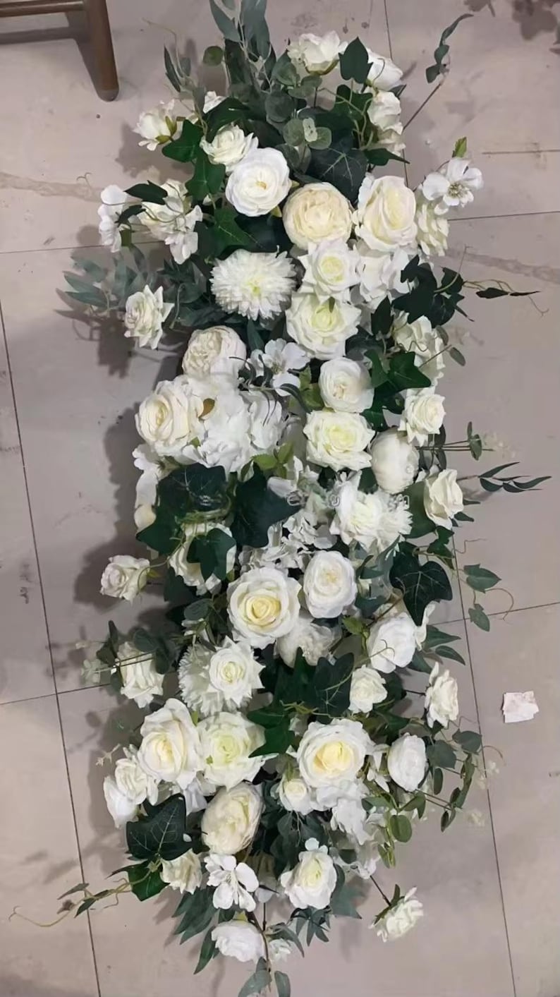 Puede incluir: Un arreglo floral largo y rectangular con rosas blancas, peon&iacute;as y otras flores blancas con follaje verde. El arreglo se coloca sobre un fondo neutro, creando una est&eacute;tica limpia y elegante.