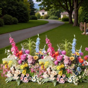 Könnte beinhalten: Ein lebendiges Blumenarrangement mit einer Vielzahl farbenfroher Blüten, darunter rosa, gelbe, blaue und orangefarbene Blumen. Die Anordnung befindet sich auf einer grünen Wiese, mit einer kurvenreichen Straße und Häusern im Hintergrund.