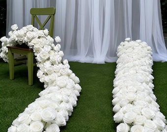 Puede incluir: Dos largos y densos caminos de flores compuestos por numerosas rosas blancas. Un camino cae en cascada desde el asiento de una silla de madera verde, mientras que el otro descansa directamente sobre el suelo. Ambos arreglos presentan rosas blancas muy juntas.