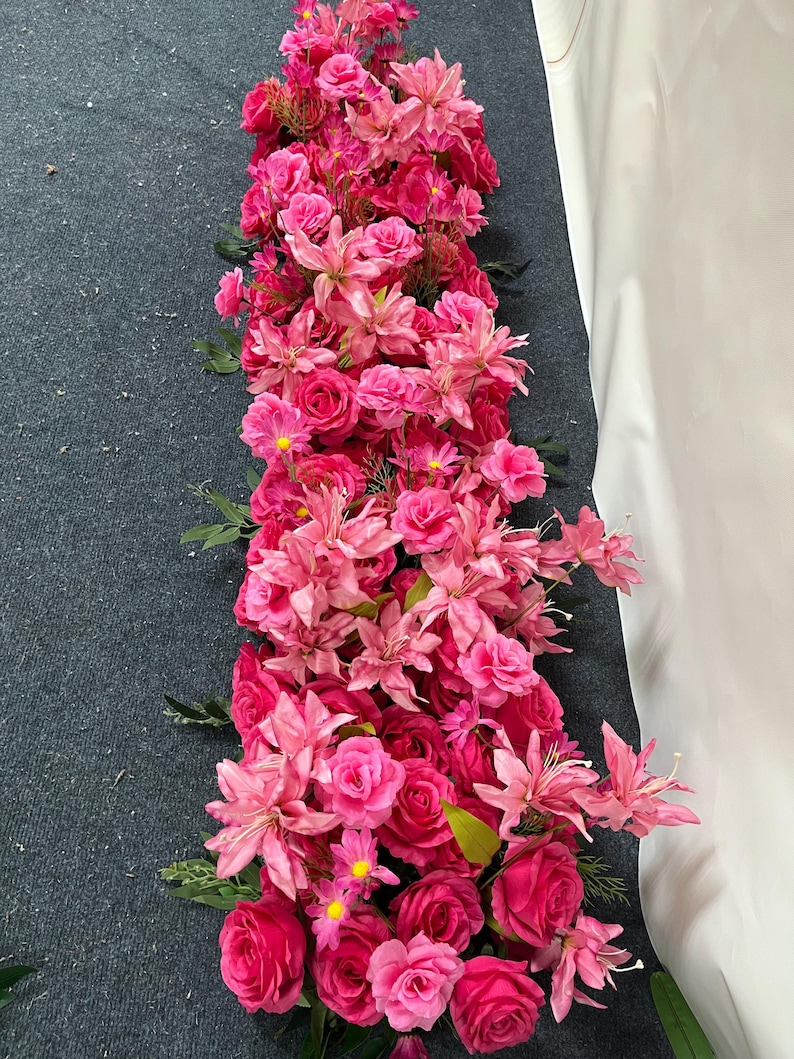 Puede incluir: Un vibrante arreglo de flores artificiales rosas y fucsias, incluyendo rosas y lirios, dispuesto en forma rectangular alargada. Las flores est&aacute;n densamente empaquetadas, creando una apariencia completa y exuberante. Hojas verdes se intercalan.