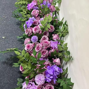 Puede incluir: Una guirnalda floral decorativa compuesta por rosas rosas, hortensias moradas y hojas verdes. El arreglo presenta una mezcla de texturas y colores, con un enfoque en las flores rosas y moradas. Ideal para bodas u otros eventos festivos.