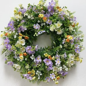 Könnte beinhalten: Ein Frühlingskranz mit einer Auswahl an lila, weißen und gelben Blumen. Der Kranz ist aus künstlichen Blumen und Grünpflanzen gefertigt.