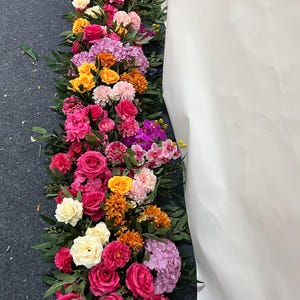 Pode incluir: Um arranjo floral vibrante com rosas, orquídeas e dálias em tons de rosa, laranja, amarelo e roxo, com folhagem verde. Ideal para casamentos ou eventos especiais. A disposição é alongada.