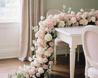 May include: A white table adorned with a cascading floral arrangement of pink roses, white hydrangeas, and delicate white filler flowers. The arrangement spills onto a wooden floor. Two pink chairs are visible.