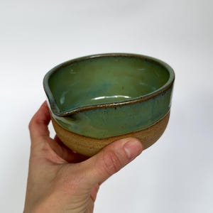 May include: A small, green ceramic bowl with a pouring spout. The bowl has a light brown rim and a slightly textured base.