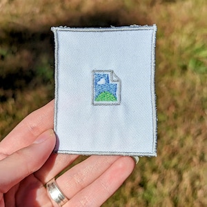 May include: White embroidered patch with a gray outline of a file icon. The file icon contains a blue sky with a white cloud and a green hill.