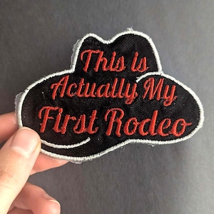 Puede incluir: Parche de hierro negro bordado en forma de sombrero de vaquero con costuras rojas. El texto dice "This is Actually My First Rodeo".