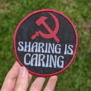 以下が含まれることがあります： 赤い鎌とハンマーのシンボルと「SHARING IS CARING」という白い文字が入った丸い黒いパッチ。パッチには赤い縁取りがあり、緑色の背景に配置されています。