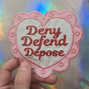 Puede incluir: Parche bordado en forma de corazón rosa con las palabras "Deny Defend Depose" en hilo rojo.