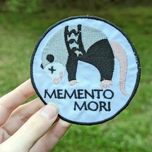 Könnte beinhalten: Ein weißer, runder gestickter Aufnäher mit einer Illustration eines schwarzen und weißen Opossums und dem Text "Memento Mori".