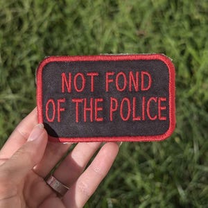 May include: A rectangular black patch with red embroidered text that reads "NOT FOND OF THE POLICE." The patch has a red border and is held against a background of green grass.