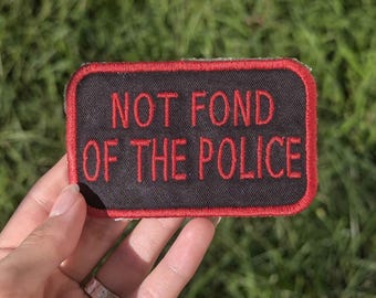 Może przedstawiać: Prostokątna czarna naszywka z czerwonym haftowanym napisem "NOT FOND OF THE POLICE". Naszywka ma czerwoną obwódkę. Naszywka jest prezentowana na tle zielonej trawy.
