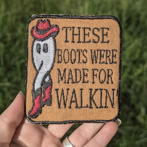 Könnte beinhalten: Gestickter Aufnäher mit beigefarbenem Hintergrund und einem Cartoon-Geist mit rotem Cowboyhut und Stiefeln. Der Text "THESE BOOTS WERE MADE FOR WALKIN" ist in schwarzen Buchstaben.