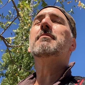 Pode incluir: Um homem com barba grisalha e um chapéu castanho olha para o céu. Ele está vestindo uma jaqueta preta e castanha com bordados dourados.