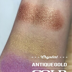 May include: Close-up of four eyeshadow swatches on a person's arm. The swatches are labeled "Crystal", "Antique Gold", "Gold", and "Violet". The colors are a light shimmery white, a copper shimmer, a gold shimmer, and a purple shimmer.