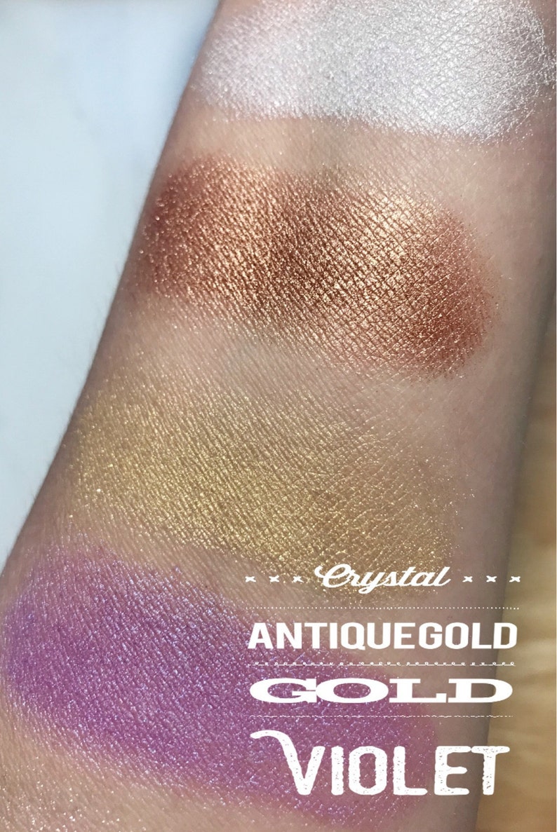 May include: Close-up of four eyeshadow swatches on a person's arm. The swatches are labeled "Crystal", "Antique Gold", "Gold", and "Violet". The colors are a light shimmery white, a copper shimmer, a gold shimmer, and a purple shimmer.