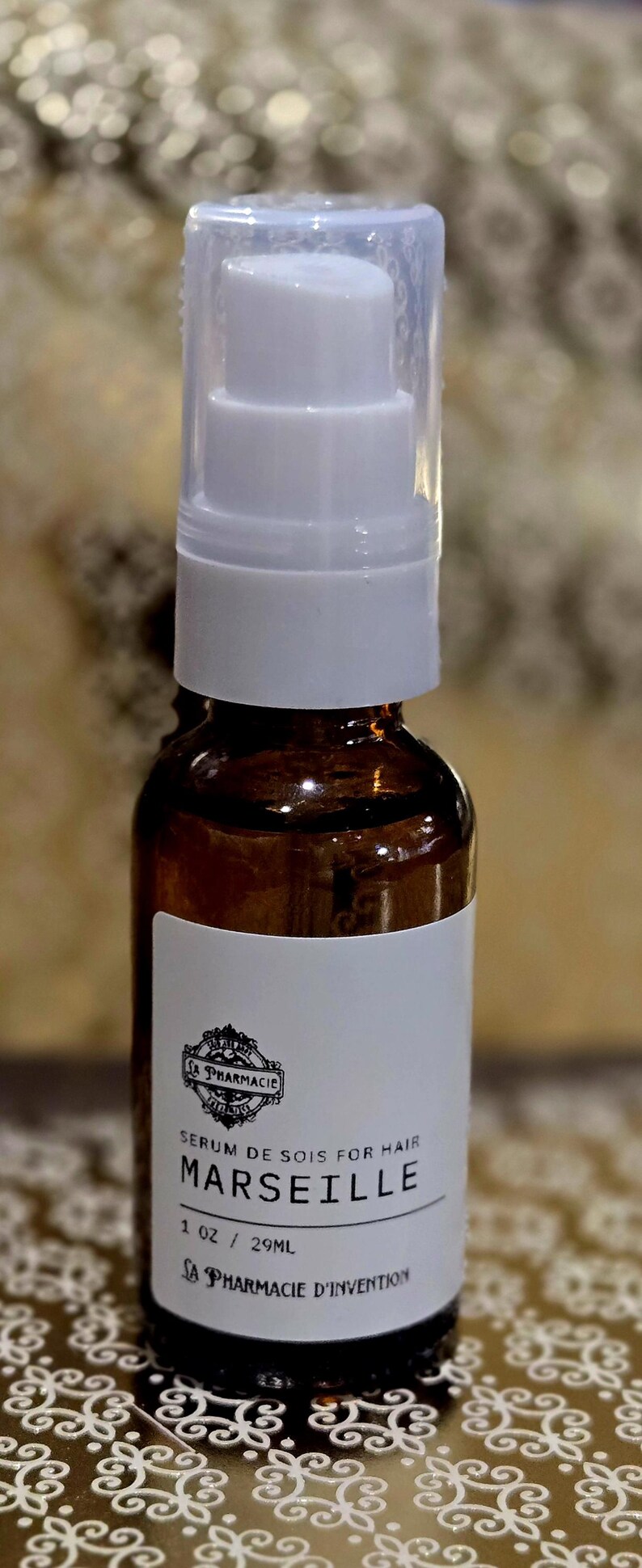 May include: A brown glass bottle with a white pump top and label. The label says "Serum de Sois for Hair Marseille 1 oz / 29mL La Pharmacie d'Invention".