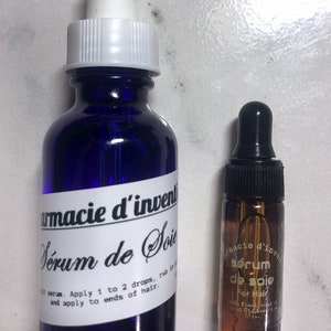 May include: Two bottles of hair serum, one blue and one amber, with dropper tops. The blue bottle has a white label with the text "armacie d'invent S&eacute;rum de Soie" and instructions for use. The amber bottle has a white label with the text "s&eacute;rum de soie For Hair".