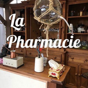 May include: A wooden cabinet with shelves filled with bottles and jars. The cabinet is labeled "La Pharmacie". A large glass bottle hangs from the ceiling and is surrounded by rope.