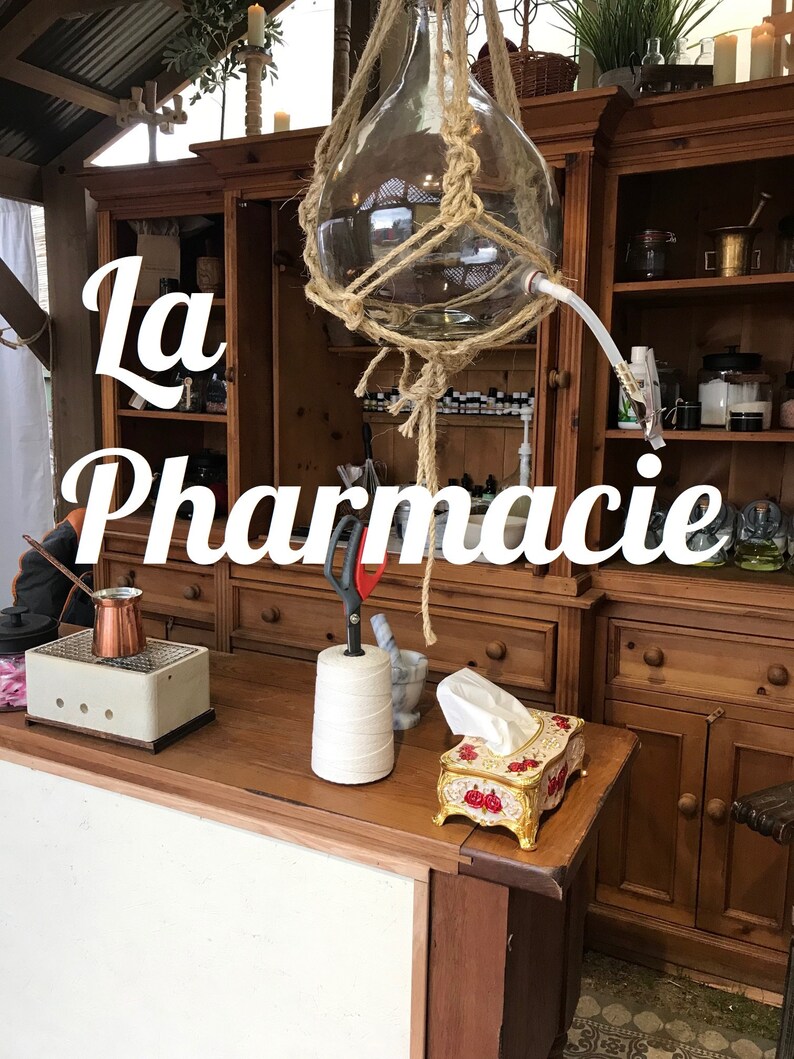 May include: A wooden cabinet with shelves filled with bottles and jars. The cabinet is labeled "La Pharmacie". A large glass bottle hangs from the ceiling and is surrounded by rope.