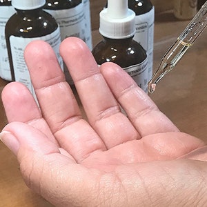 Puede incluir: Una mano con un frasco gotero de vidrio transparente con una tapa blanca y un l&iacute;quido marr&oacute;n en su interior. El frasco est&aacute; etiquetado con "Farmacia di"