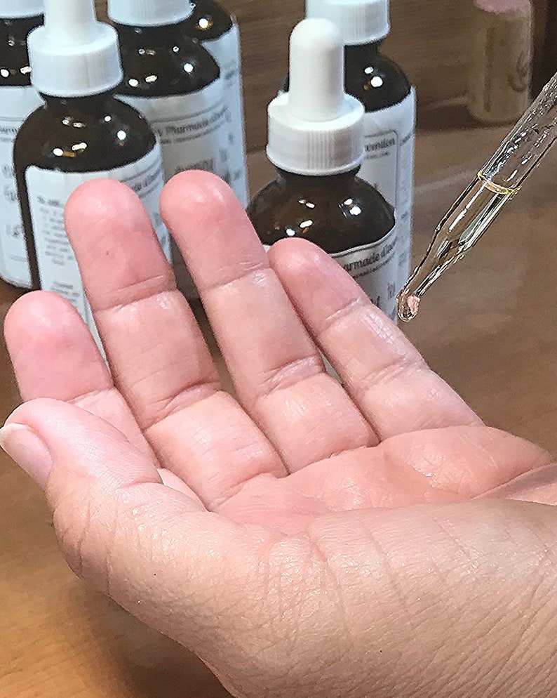 Puede incluir: Una mano con un frasco gotero de vidrio transparente con una tapa blanca y un l&iacute;quido marr&oacute;n en su interior. El frasco est&aacute; etiquetado con "Farmacia di"