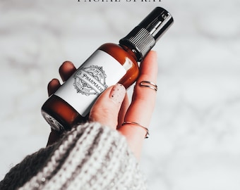 May include: A brown glass bottle with a black spray nozzle, labeled "LA PHARMACIE D'INVENTION GREEN TEA MIST FACIAL SPRAY". The bottle is held in a hand wearing a gray sweater.