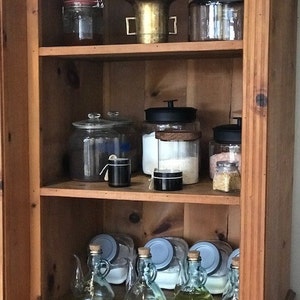 May include: A wooden cabinet with three shelves filled with glass jars and bottles. The jars and bottles are filled with various items, including spices, grains, and liquids. There is also a mortar and pestle on the top shelf.