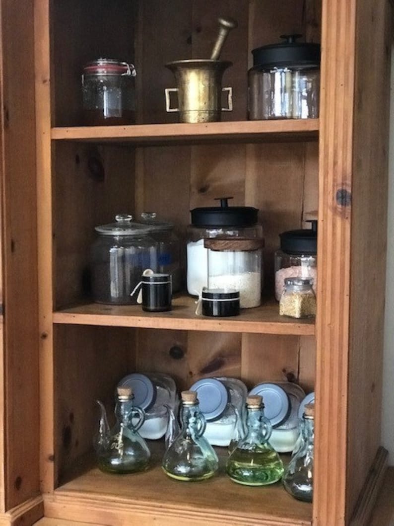 May include: A wooden cabinet with three shelves filled with glass jars and bottles. The jars and bottles are filled with various items, including spices, grains, and liquids. There is also a mortar and pestle on the top shelf.