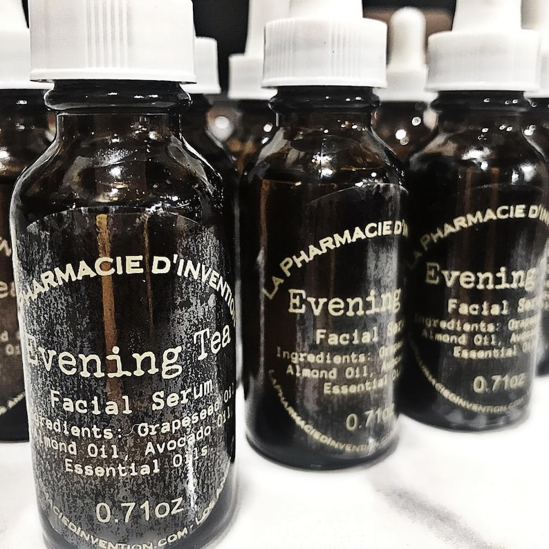 Puede incluir: Una colecci&oacute;n de botellas de vidrio &aacute;mbar con goteros blancos y etiquetas que dicen "La Pharmacie d'Invention" y "Evening Facial Serum" o "Evening Tea Facial Serum". Las etiquetas enumeran los ingredientes, que incluyen aceite de semilla de uva, aceite de almendras, aceite de aguacate y aceites esenciales. Las botellas son de 0,71 oz.