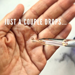May include: A hand with a few drops of clear liquid on it. A glass dropper with a gold tip is dispensing the liquid. The text "JUST A COUPLE DROPS..." is visible above the hand.