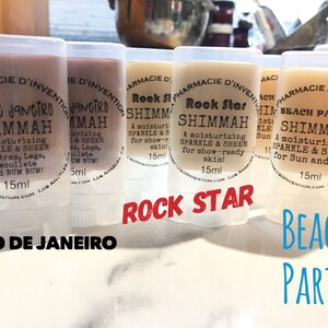Könnte beinhalten: Sechs kleine, transparente Plastikröhrchen mit weißen Etiketten und schwarzem Text. Die Röhrchen enthalten eine schimmernde Lotion. Die Etiketten lauten "Rio de Janeiro Shimma, Rock Star Shimma, Beach Party Shimma".